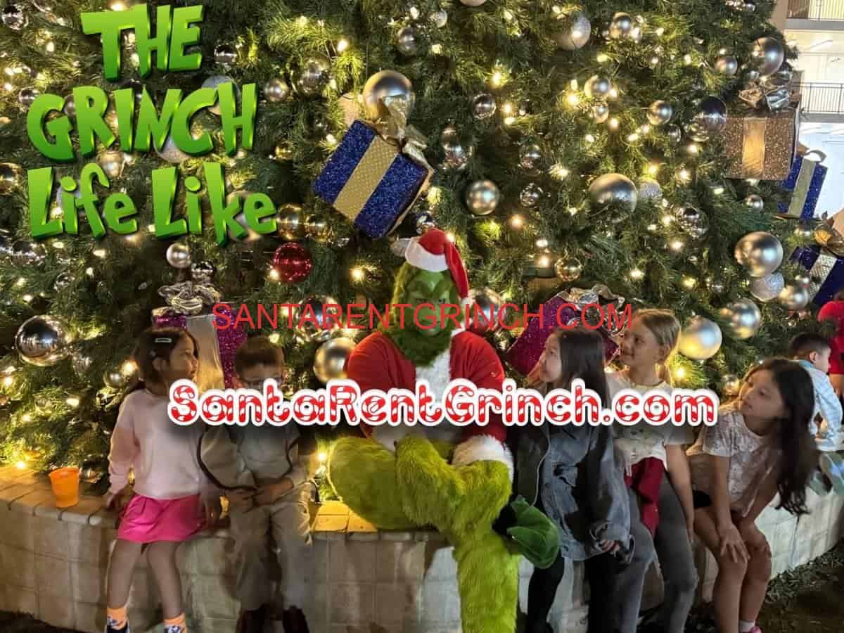 Grinch sitting with a group of children in front of a giant ornament-covered Christmas tree